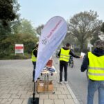 Stadt-Rosenheim-Fahne und Mitarbeitende bei der Übergabe von Brezen an Radfahrende in der Küpferlingstraße.