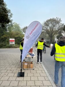 Stadt-Rosenheim-Fahne und Mitarbeitende bei der Übergabe von Brezen an Radfahrende in der Küpferlingstraße.