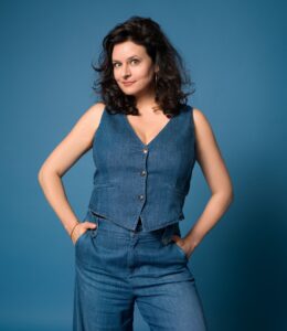 3: Angela Ascher in Jeansweste und -hose vor blauem Hintergrund.