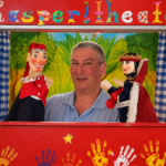 Ein Spieler mit Kasperl- und Märchenkönig-Handpuppen vor roter Bühnenkulisse.