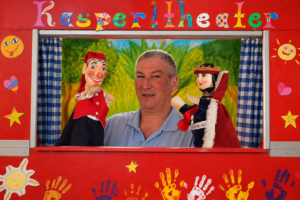 Ein Spieler mit Kasperl- und Märchenkönig-Handpuppen vor roter Bühnenkulisse.