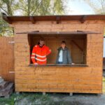 Mitarbeiter des Bauhofs und eine Mitarbeiterin der Tourist‑Info stehen in einer reparierten Weihnachtsmarkthütte.
