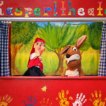 Kasperl und Mucki vor der roten Bühnenkulisse mit Kinderhandabdrücken.