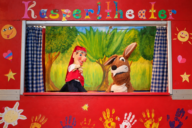 Kasperl und Mucki vor der roten Bühnenkulisse mit Kinderhandabdrücken.