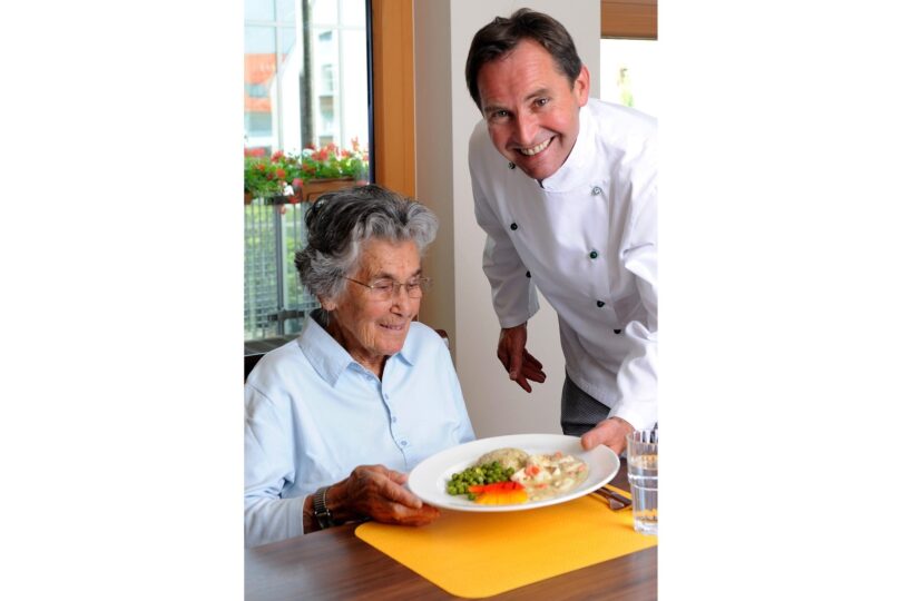 Koch serviert einer Seniorin ein Essen in einer Einrichtung