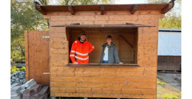 Mitarbeiter des Bauhofs und eine Mitarbeiterin der Tourist‑Info stehen in einer reparierten Weihnachtsmarkthütte.