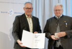 Zwei Männer sind festlich gekleidet. Sie halten Urkunde und Medaille in den Händen.