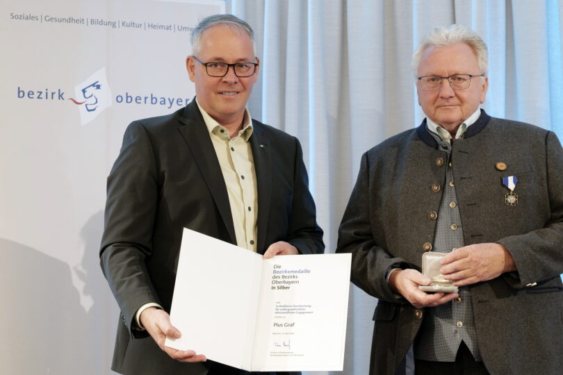 Zwei Männer sind festlich gekleidet. Sie halten Urkunde und Medaille in den Händen.