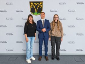 Drei Personen stehen vor einer Wand mit dem Schriftzug der Großen Kreisstadt Traunstein.