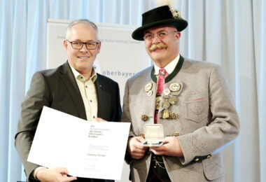 Zwei Männer sind festlich gekleidet. Sie halten Urkunde und Medaille in den Händen.