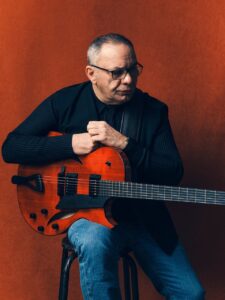 Bireli Lagrene mit Gitarre vor rotem Hintergrund.