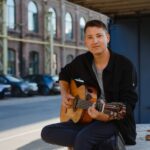 Daniel Stelter spielt Gitarre vor einem Backsteingebäude.