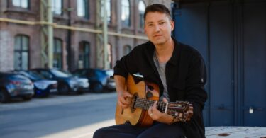 Daniel Stelter spielt Gitarre vor einem Backsteingebäude.
