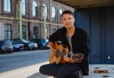 Daniel Stelter spielt Gitarre vor einem Backsteingebäude.