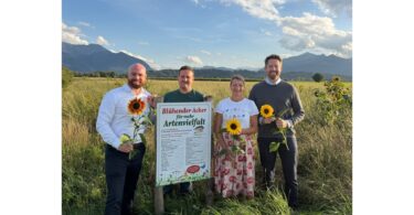 Personen mit Sonnenblumen neben einem Schild 'Blühender Acker für mehr Artenvielfalt'.