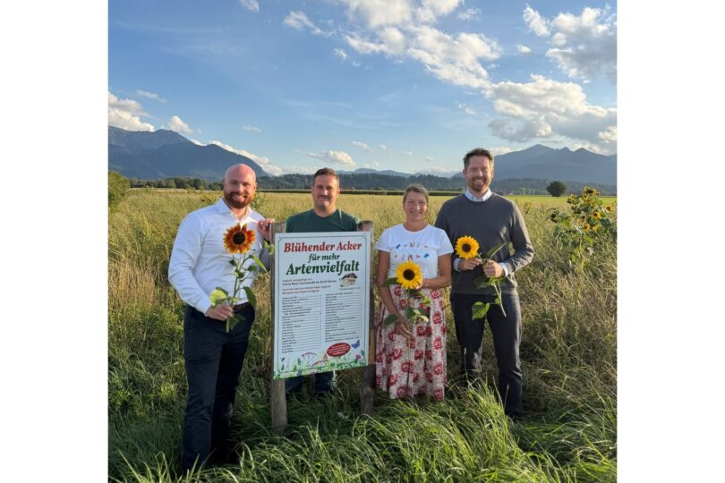 Personen mit Sonnenblumen neben einem Schild 'Blühender Acker für mehr Artenvielfalt'.