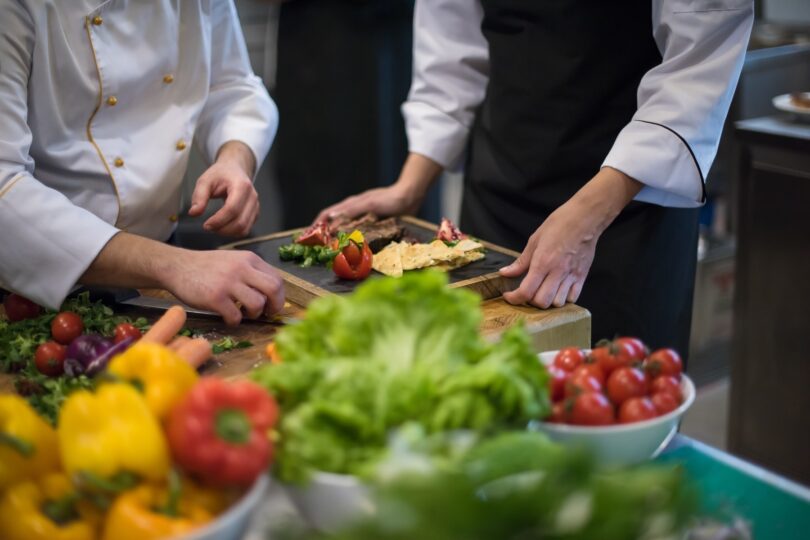 Köche richten Speisen an, frisches Gemüse liegt im Vordergrund.